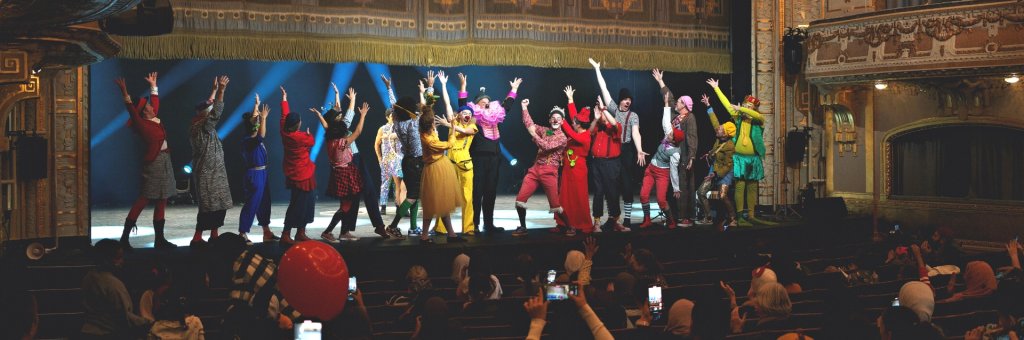 Många artister i färgglada kläder står på Dramatens stora scen med armarna i luften framför publiken där många håller upp mobilen för att filma