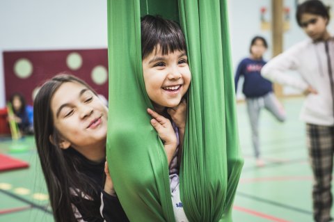 Ett barn sitter i ett grönt tyg som hänger från taket och ett annat barn står bakom. Båda skrattar.