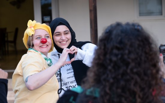 En clown med röd näsa och en flicka i hijab formar ett hjärta tillsammans med varsin hand