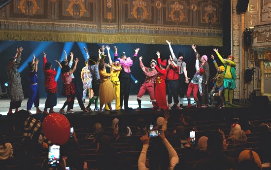 Många artister i färgglada kläder står på Dramatens stora scen med armarna i luften framför publiken där många håller upp mobilen för att filma